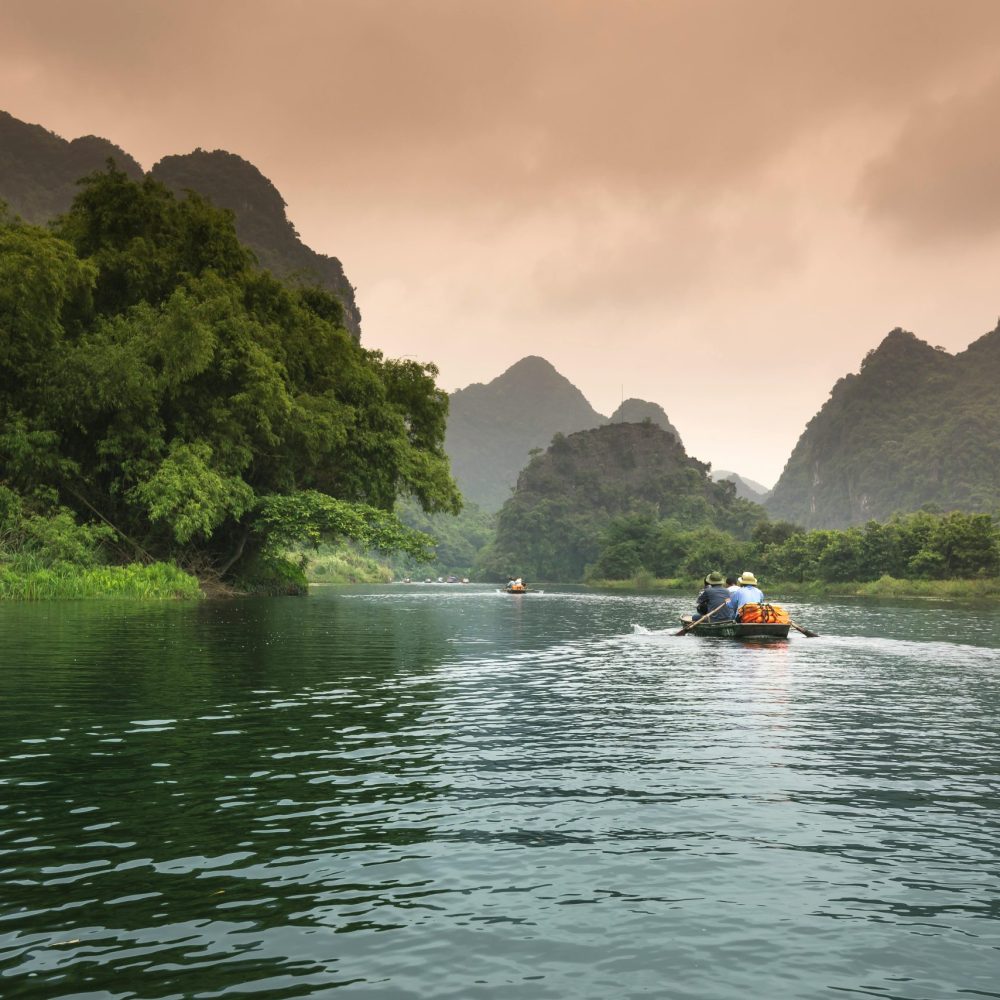 Two people rowing a boat through a serene river surrounded by lush greenery and mountains.