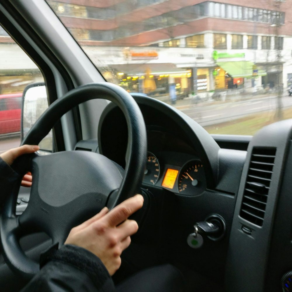 Hands on a steering wheel driving a van through an urban area with shops visible outside.